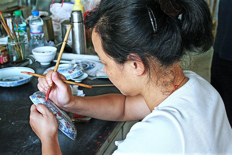 Eine Arbeiterin bei der Aufglasurmalerei in der früheren Jiayang–Fabrik. Sie hält und führt dabei mehrere Pinsel verschiedener Größen, die sie wechselweise einsetzt, um komplexe Dekors umzusetzen.